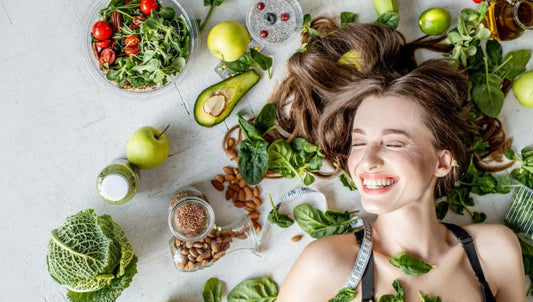 Foto de cima de mulher deita sorrindo com cabelo espalhado e diversos vegetais, frutas, grãos ao seu redor